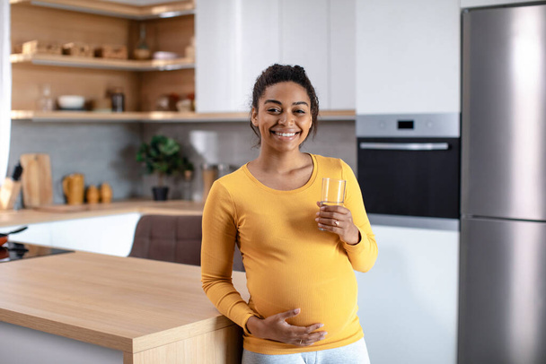 Щаслива молода вагітна афро-американська жінка торкається великого живота і тримає склянку води в мінімалістському кухонному інтер'єрі, вільному просторі. Баланс з аквою, домашня охорона здоров "я, харчування та корисне харчування. - Фото, зображення