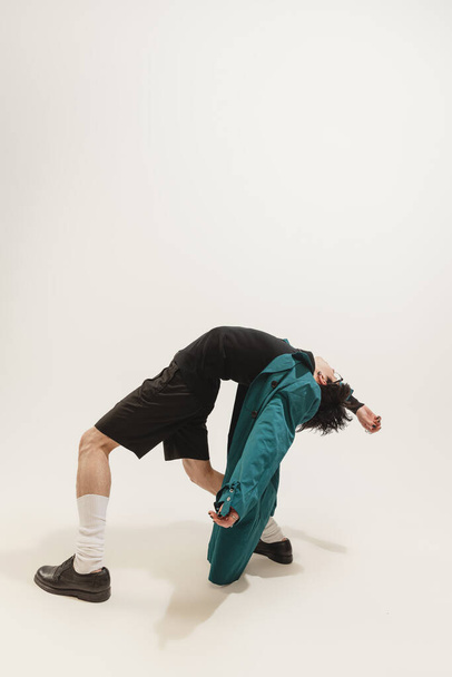 Portrait of stylish young man in black outfit and green coat posing isolated over grey studio background. Expressive dance. Concept of modern fashion, art photography, style, queer, uniqueness, ad - Φωτογραφία, εικόνα