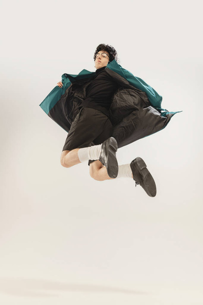 Portrait of stylish young man in black outfit and green coat posing, jumping isolated over grey studio background. Concept of modern fashion, art photography, style, queer, uniqueness, ad - Foto, Imagen