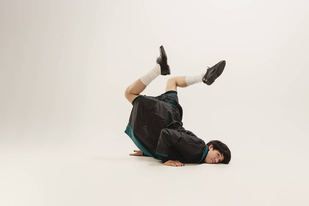 Stylish young man in black outfit and green coat posing, lying on floor isolated over grey studio background. Dance.Concept of modern fashion, art photography, style, queer, uniqueness, ad - Foto, Imagen