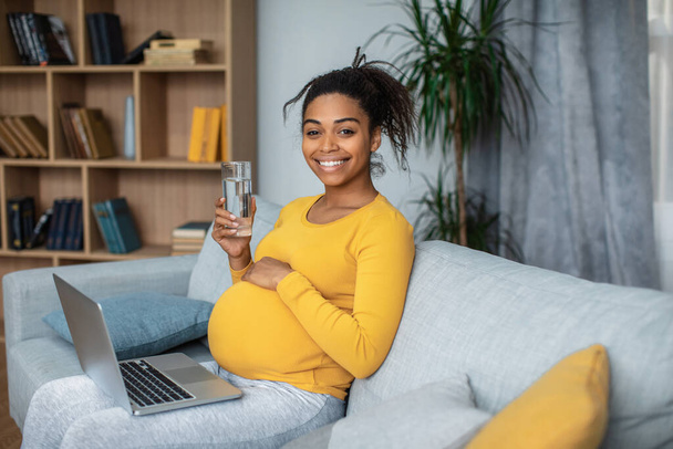Happy young pregnant african american female touches big belly, drinks water and watches video on laptop in living room interior. Work, business and study during pregnancy, motherhood and online chat - 写真・画像