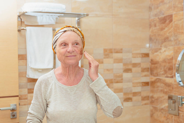 Mature woman standing at the mirror in the bathroom and putting a scrub on her face. Elderly woman taking care of her face - Foto, immagini