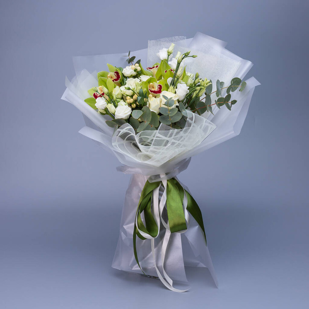 Flower bouquet in light green color, which is wrapped in light floral paper stands on a blue background. Delicate bouquet of white roses, eustoma, green irises and eucalyptus twigs. - Photo, image
