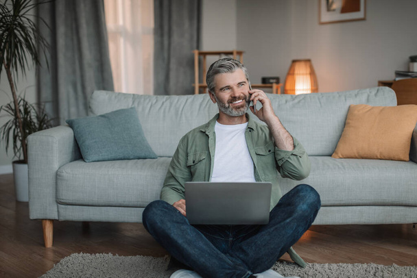 Smiling caucasian retired male with beard sits on floor with laptop, calls by phone in living room interior. Pensioner work at home, freelance, work and business remotely, during covid-19 outbreak - 写真・画像