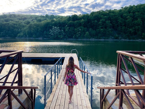 Вид сзади на женщину в летнем платье, стоящую на деревянном понтоне возле озера - Фото, изображение