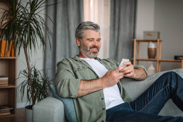 Улыбающийся кавказский взрослый мужчина с бородой смотрит видео, играет в игру на смартфоне в интерьере гостиной. Пенсионер весело провести время в свободное время, болтая онлайн и отдохнуть с устройством связи covid-19 пандемии - Фото, изображение