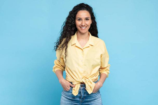Happy Middle Eastern Lady With Long Brunette Hair Smiling To Camera Posing Holding Hands In Pockets, Wearing Casual Clothes Over Blue Background. Woman Standing In Studio - Fotó, kép
