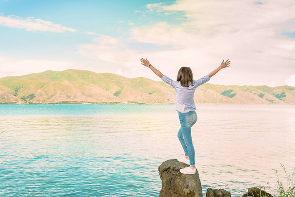 Een jong meisje in een blauwe blouse staat op de stenen oever van het meer en spreidt haar armen uit. - Foto, afbeelding