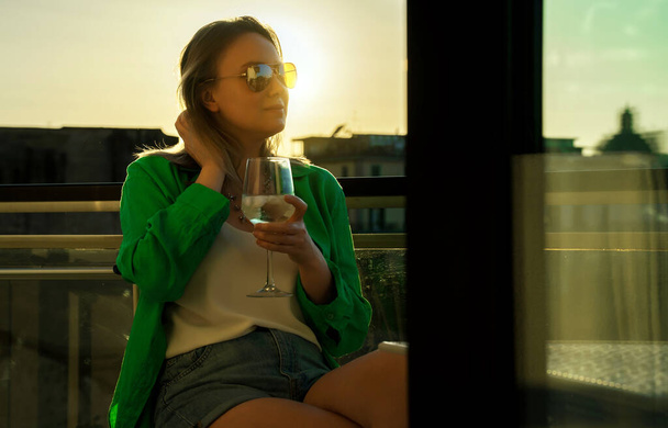 Γυναίκα με λευκό κρασί απολαμβάνει το ηλιοβασίλεμα στο μπαλκόνι της. - Φωτογραφία, εικόνα