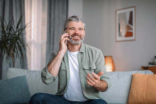Cheerful caucasian senior male with beard calls by phone in living room interior. Retired talking to client at home, communication and social distance, new normal and health care due covid-19 outbreak - Fotografie, Obrázek