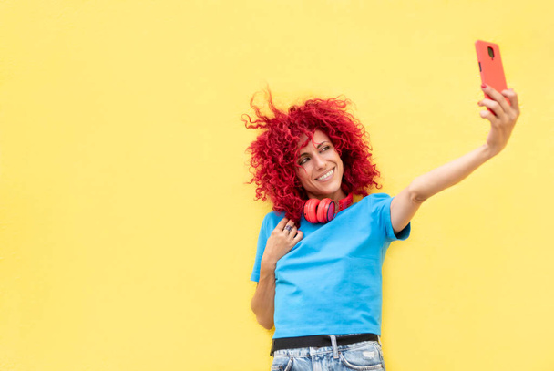 портрет усміхненої латинки з рудим афро-волоссям, що приймає селфі на жовтому тлі, одягнений у синю футболку та червоні навушники на шиї
. - Фото, зображення