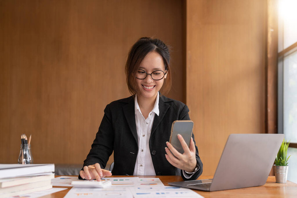 Asian woman holding smart phone while using calculator for business financial accounting calculate money bank loan rent payments manage expenses finances taxes doing paperwork concept, close up - 写真・画像