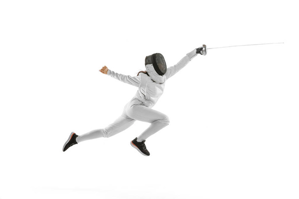 Dynamic portrait of female fencer in sports costume and fencing mask holding rapier in hand and training isolated on white background. Sport, youth, healthy lifestyle, achievements. Copy space for ad - Foto, Imagem