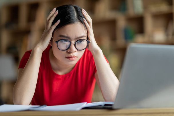Skoncentrowany zajęty młoda Azjatka dziewczyna w okularach trzymać głowę z rękami, spojrzeć na komputer w pokoju wnętrza. Problemy w nauce zdalnie, praca domowa, odległość społeczna, przygotowanie do egzaminu w domu i znaleźć błąd - Zdjęcie, obraz