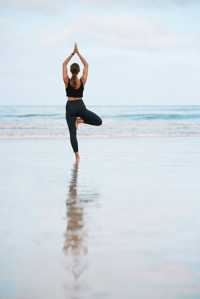 Rearview shot van een jonge vrouw doen een boom pose tijdens het beoefenen van yoga op het strand. - Foto, afbeelding