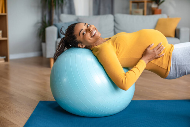 Joyeux heureux jeune femme enceinte afro-américaine toucher gros ventre, faire des exercices pour le dos sur ballon en forme dans le salon intérieur, espace libre. Soins corporels pendant la grossesse, sports à la maison et entraînement - Photo, image
