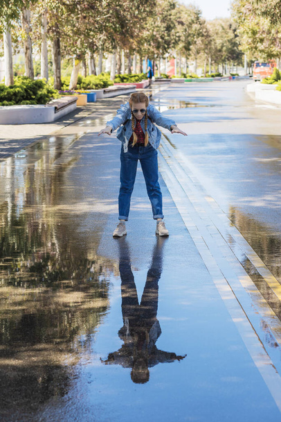 Kot elbiseli bir kadın, vücudu eğik ve kollarını bir parktaki ıslak bir sokakta ileri doğru uzatarak duruyor. Yağmurdan sonra yürü. Neşeli imaj. - Fotoğraf, Görsel