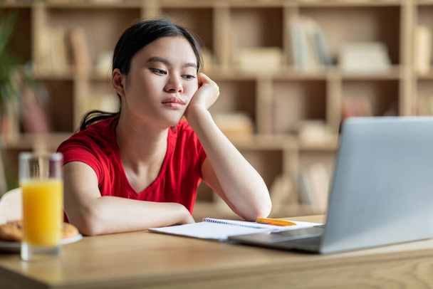 Üzgün, Asyalı kız öğrenci, içerideki bilgisayardan video dersi izliyor. Eğitim ve öğrenim sorunları, uzaktan eğitim, sosyal mesafe, sınav hazırlığı ve evde covid-19 salgını nedeniyle sıkıcılık - Fotoğraf, Görsel