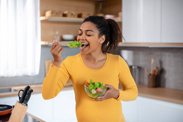 Усміхаючись голодна молода вагітна афроамериканка їсть салат з органічних овочів в мінімалістичному кухонному інтер'єрі. Правильне харчування, охорона здоров'я, вегетаріанство та дієта під час очікуваної дитини
 - Фото, зображення