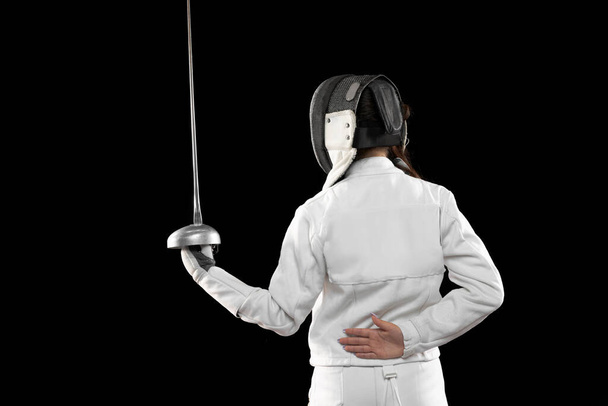 Defender moving. One sportsman, female fencer in white fencing costume in basic stance isolated on black background. Sport, youth, hobbies, achievements, goal. Girl practicing with rapier - Photo, image