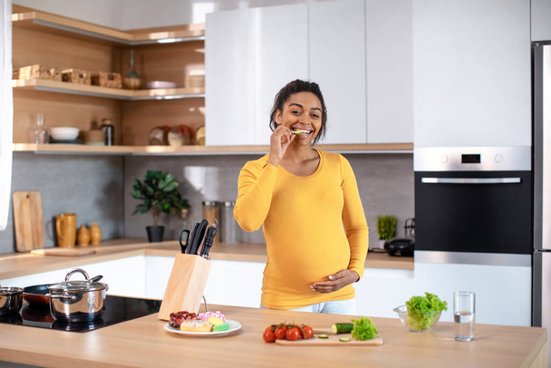 Cheerful young pregnant african american female touching big belly, call by phone at table with sweets, vegetables and water in minimalist kitchen interior. Health care, diet and useful nutrition - Foto, Bild