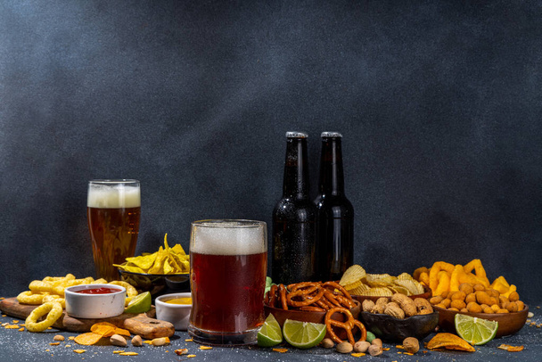 Beer with various salted snacks set. Black table background with traditional party snacks, beer bottles and glasses, with chips, onion rings, salted nuts, crisps and sauces top view copy space - Fotografie, Obrázek