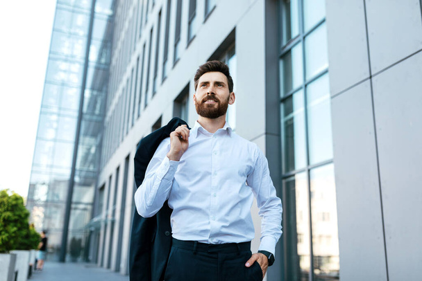 Confident handsome european millennial bearded businessman with jacket on shoulder near modern office building, outdoor, free space. Business, successful ceo manager, boss and professional work - Photo, Image