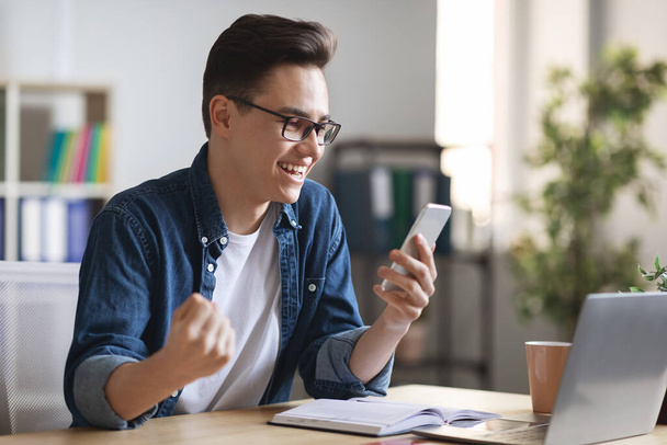 Good News. Joyful Young Man Celebrating Success With Smartphone At Workplace In Office, Millennial Male Employee Sitting At Desk And Looking At Cellphone Screen With Excitement, Shaking Fist With Joy - Foto, imagen