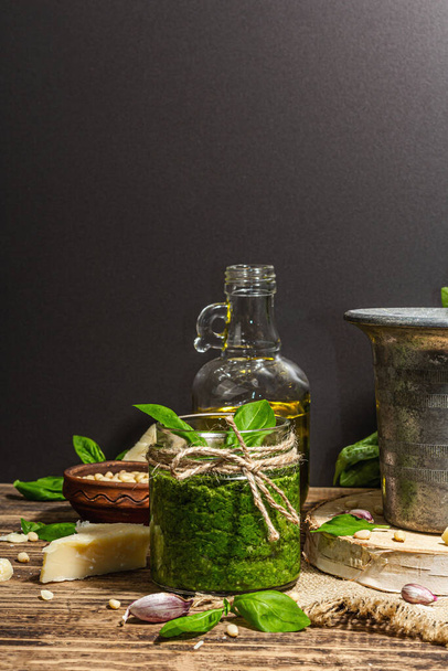 Homemade Italian basil pesto sauce in a vintage mortar with pestle. Fresh bunch of leaves, parmesan, pine nuts, and olive oil. Trendy hard light, dark shadow, old wooden background, copy space - Zdjęcie, obraz