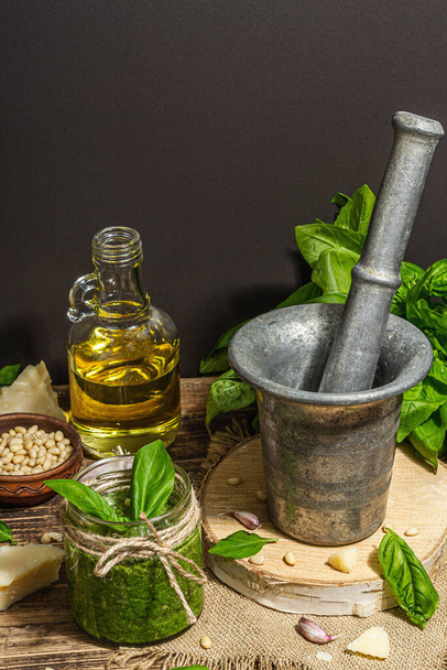 Homemade Italian basil pesto sauce in a vintage mortar with pestle. Fresh bunch of leaves, parmesan, pine nuts, and olive oil. Trendy hard light, dark shadow, old wooden background, copy space - Photo, image