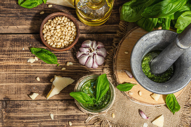 Homemade Italian basil pesto sauce in a vintage mortar with pestle. Fresh bunch of leaves, parmesan, pine nuts, and olive oil. Trendy hard light, dark shadow, old wooden background, top view - Photo, image