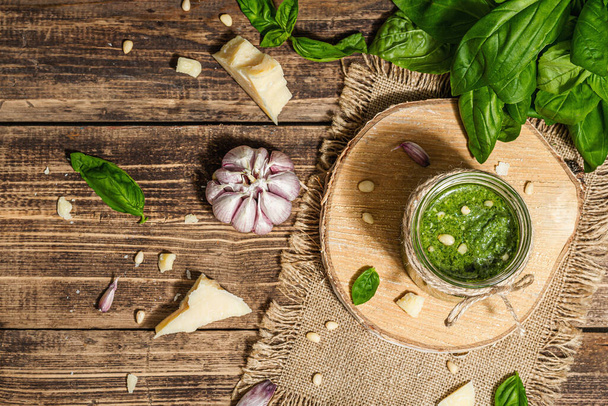 Homemade Italian basil pesto sauce in a glass jar. Fresh bunch of leaves, parmesan, pine nuts, and olive oil. Trendy hard light, dark shadow, old wooden background, top view - Photo, image