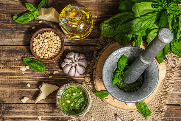 Homemade Italian basil pesto sauce in a vintage mortar with pestle. Fresh bunch of leaves, parmesan, pine nuts, and olive oil. Trendy hard light, dark shadow, old wooden background, top view - Foto, afbeelding