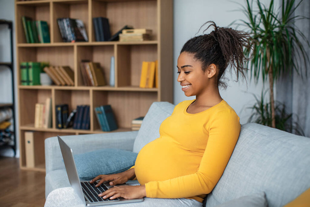 Cheerful millennial pregnant african american lady with big belly typing on laptop in living room interior. Work, study, business during pregnancy, motherhood and online chat and social distance - Photo, image