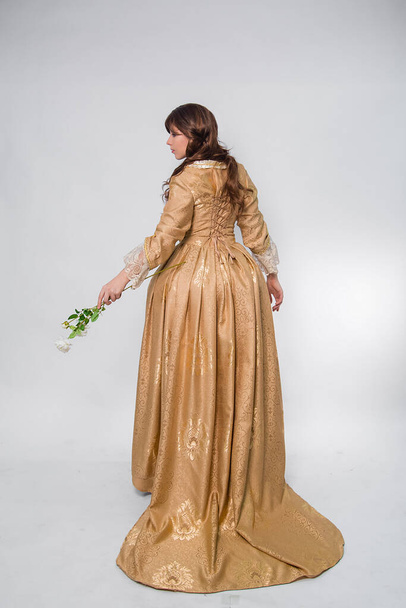 Full length portrait of a woman in a gold dress in the style of the rococo era, standing with her back forward and posing with a flower in hand isolated on a white background. - Photo, image