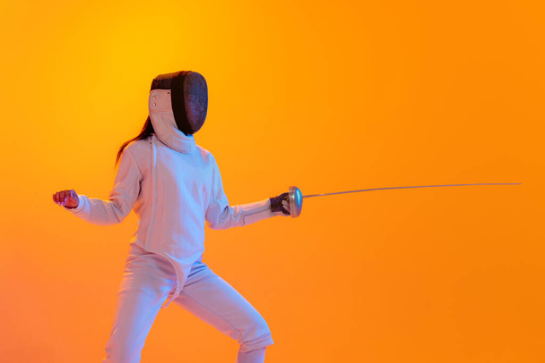 En garde. Studio shot of professional fencer in white fencing costume and mask in action, motion isolated on orange color background. Concept of sport, youth, activity, skills, achievements, ad. - Photo, Image