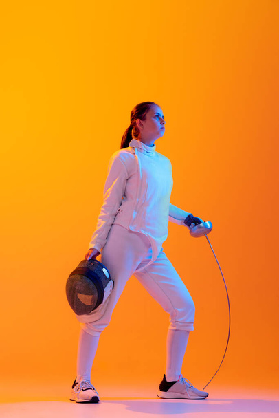 Rest time. Professional fencer in white fencing costume and mask in action, motion isolated on orange color background. Concept of sport, youth, activity, skills, achievements, ad. - Photo, image