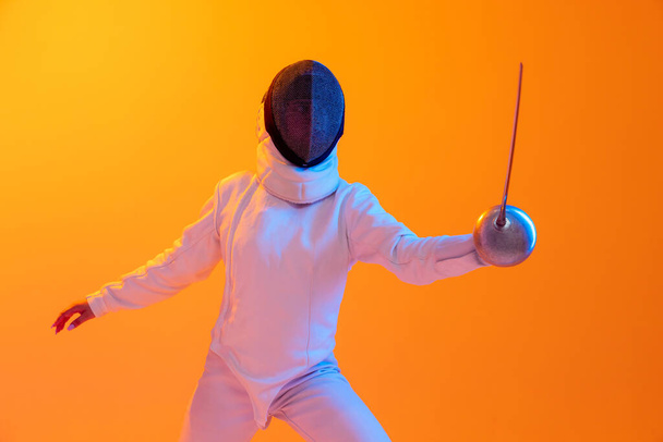 Studio shot of professional fencer in white fencing costume and mask in action, motion isolated on orange color background. Concept of sport, youth, activity, skills, achievements, ad. - Foto, Imagen