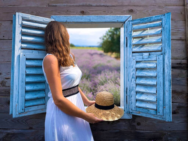 Femme en robe blanche debout près d'une fenêtre en bois à travers laquelle un champ de lavande peut être vu - Photo, image