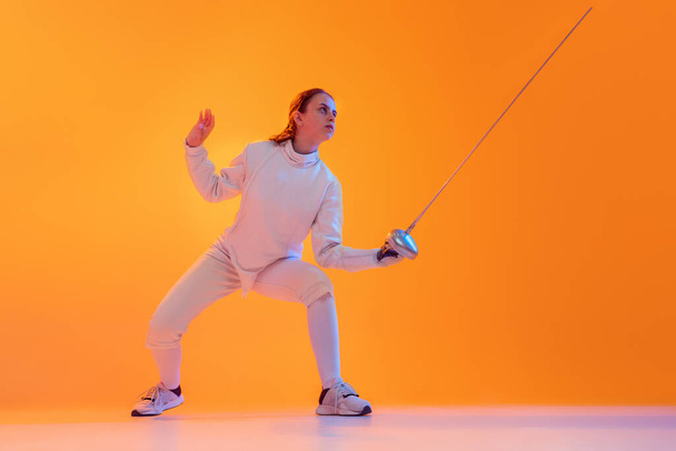 Studio shot of professional fencer in white fencing costume and mask in action, motion isolated on orange color background. Concept of sport, youth, activity, skills, achievements, ad. - Photo, image