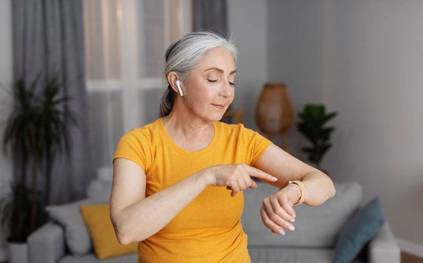 Modern senior woman in sports outfit checking smartwatch, wearing earphones while training at home in living room. Aged female using modern gadget for exercising - Foto, imagen