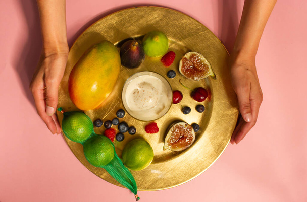 Le mani della donna servono un piatto ben servito, piatto di frutta tropicale fresca su una vista dall'alto del tavolo rosa. Ragazza senza volto che serve sul tavolo. Fichi, mango, bacche, ciliegie, lime, gelato su un vassoio - Foto, immagini