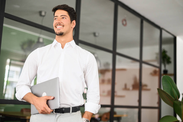 High-skilled smiling young businessman carrying laptop in a modern office, white collar worker ready for new working day, inspired with new ideas male start-up owner looking aside - Foto, imagen