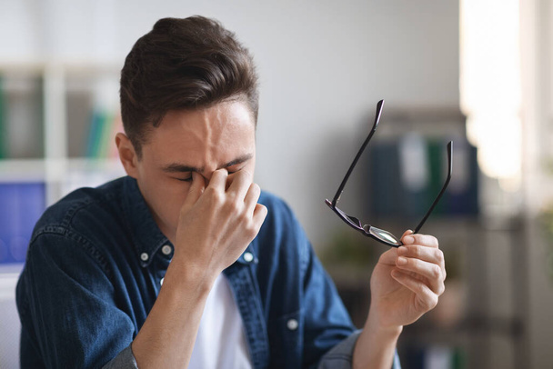 Portret van een vermoeide jongeman die glas afneemt en neusbrug wrijft, close-up shot van Millennnial Office Employee die last heeft van oogvermoeidheid op het werk na een dag hard werken, met gezondheidsproblemen - Foto, afbeelding