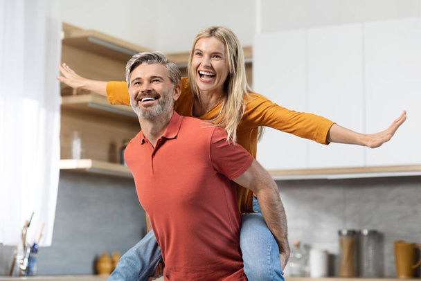 Joyful pretty blonde lady piggybacking her cheerful husband, looking at copy space and smiling, middle aged couple going to cook nice meal together, having fun, kitchen interior - Photo, Image