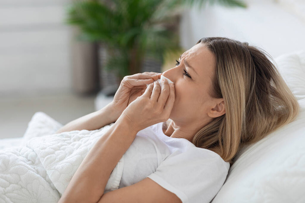 Sick millennial woman with long blonde hair lying in bed at home, sneezing nose, using tissues, suffering from runny nose while having coronavirus or seasonal flu, closeup, copy space - Photo, Image