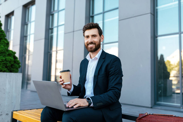 Bin yıllık Avrupa sakallı, takım elbiseli bir işadamı laptopta çalışıyor, ofis binasının yanındaki bankta kahve içiyor. İş dünyası, kahve molası, yeni normal yaşam tarzı ve dijital göçebe - Fotoğraf, Görsel