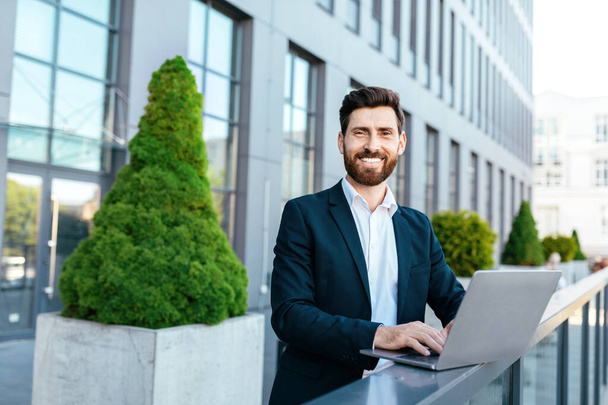 Glad confident european millennial bearded businessman in suit typing on laptop on balcony near modern office building. Work and trade in city. Business remote outdoor, new normal and digital nomad - Zdjęcie, obraz