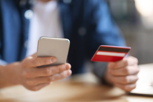 E-Commerce Concept. Closeup Shot Of Unrecognizable Male Using Smartphone And Credit Card, Cropped Image Of Young Man Sitting At Desk And Making Online Payments Or Internet Shopping Via Mobile Phone - Photo, Image