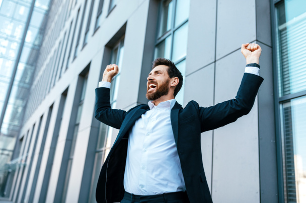 Happy satisfied excited european millennial bearded businessman in suit rejoices in successful deal or win, rises hands up near modern office building, outdoor. Manager, emotions of victory, gestures - 写真・画像